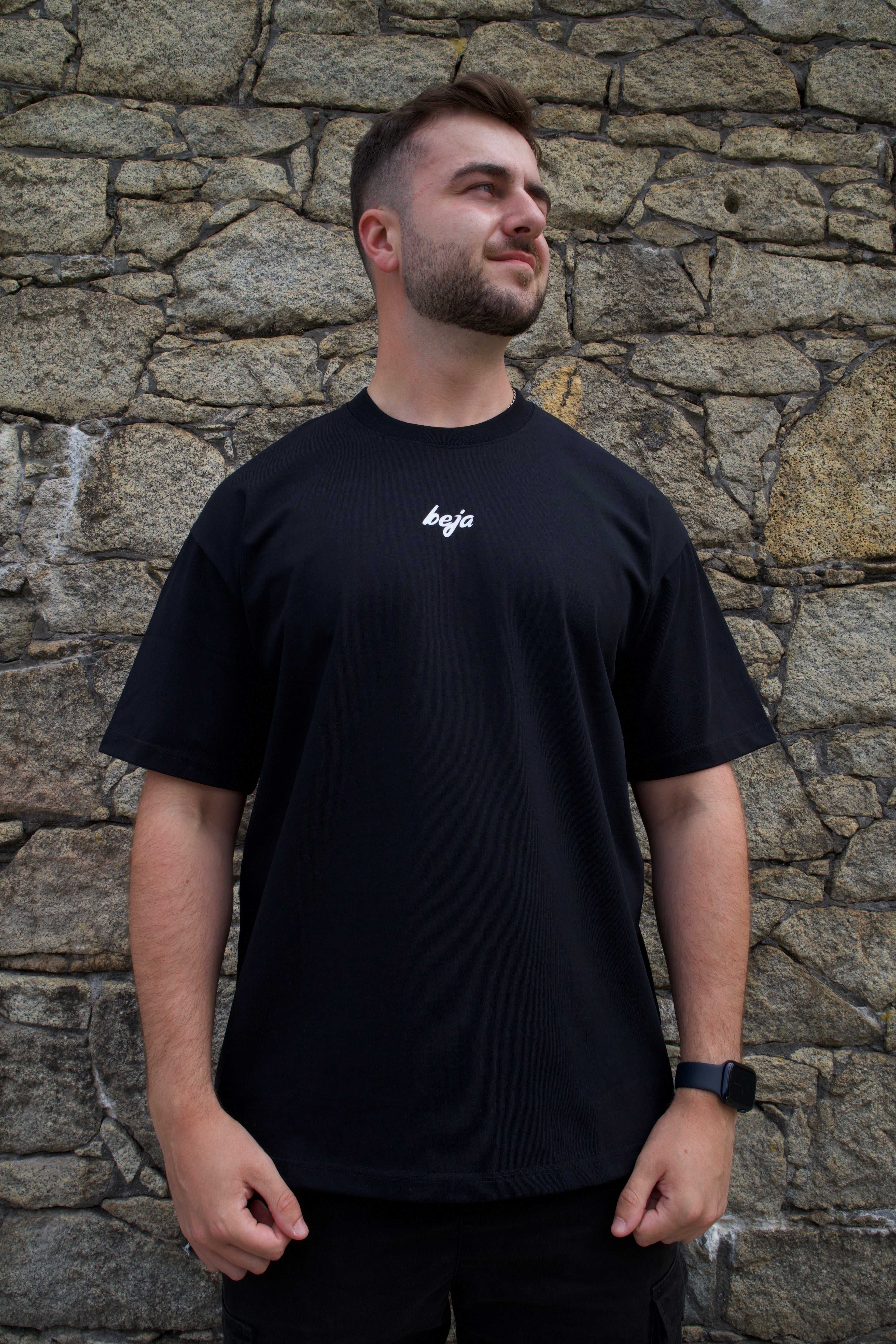 A man wearing a black short-sleeve t-shirt with a small white logo on the left chest, standing against a textured stone wall.