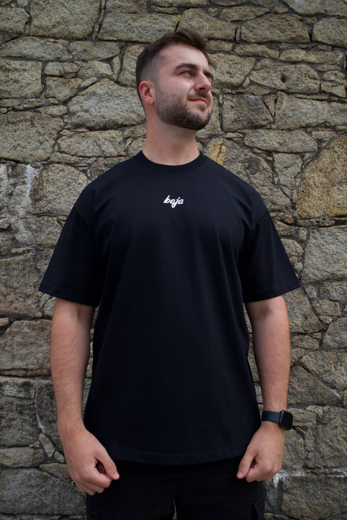 A man wearing a black short-sleeve t-shirt with a small white logo on the left chest, standing against a textured stone wall.