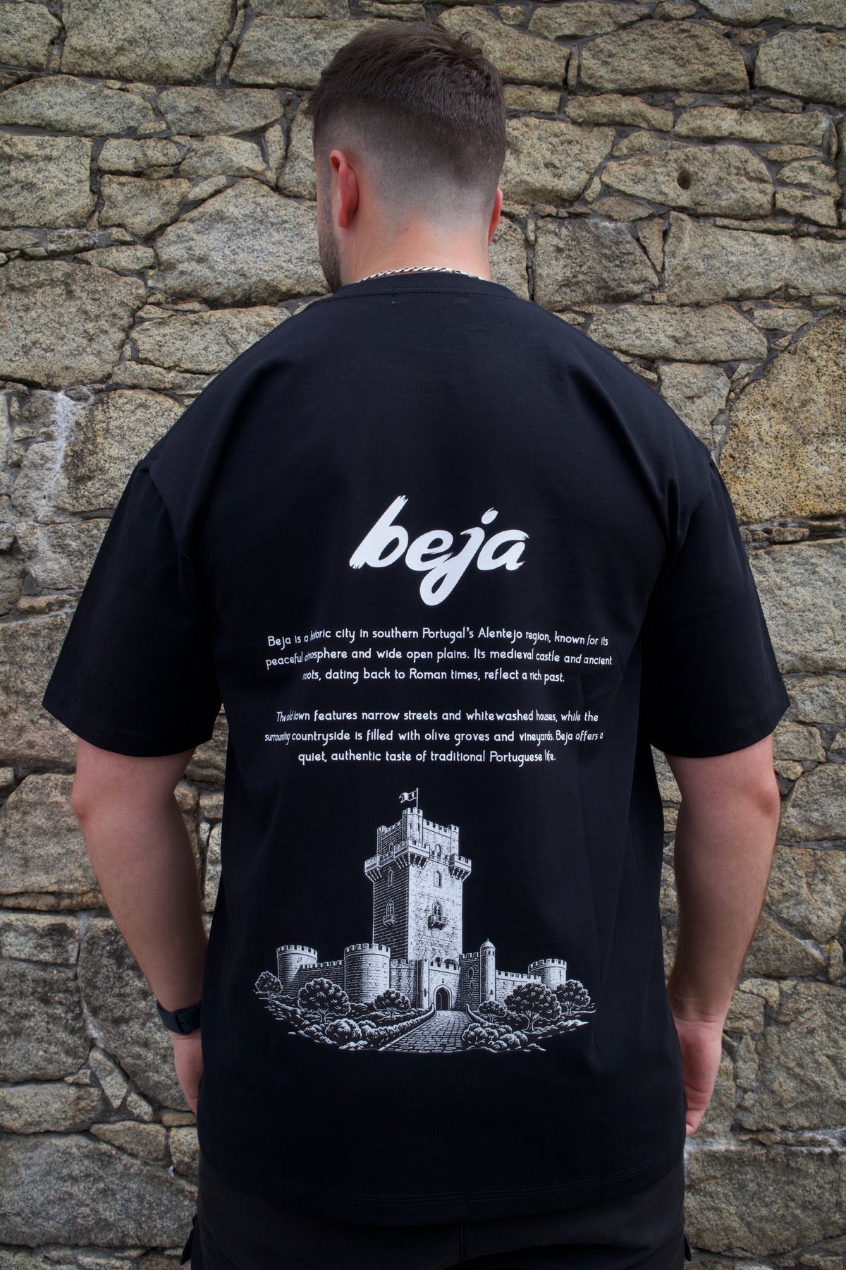 A man viewed from behind wearing a black t-shirt with a white graphic print of a castle and the word 'beja' on the back, standing against a stone wall.