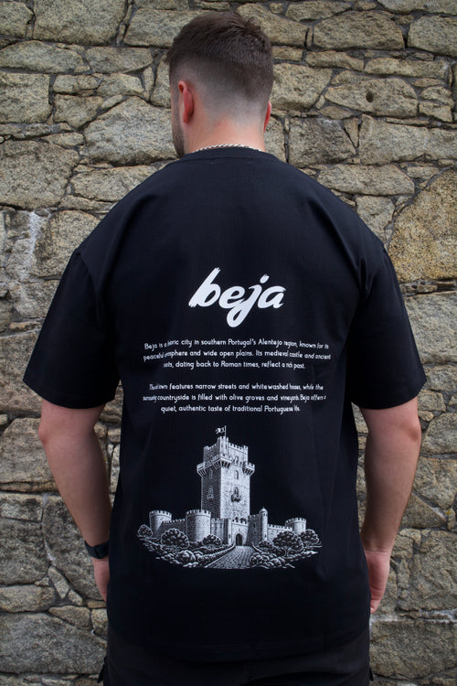 A man viewed from behind wearing a black t-shirt with a white graphic print of a castle and the word 'beja' on the back, standing against a stone wall.